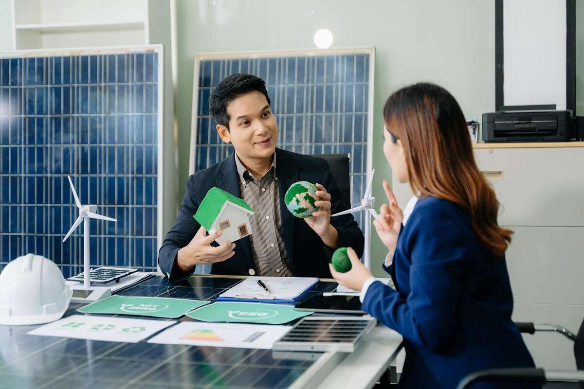 A man and a woman in business attire sitting at a desk surrounded by solar panels and miniature wind turbines, discussing environmental sustainability while holding a model house and a small globe.