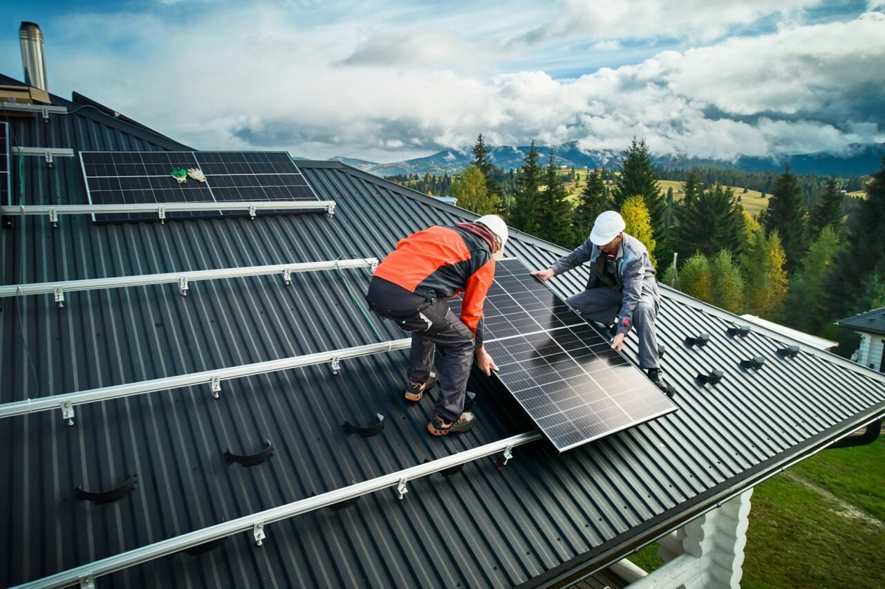 Two workers in white helmets positioning a solar panel on a black metal roof.